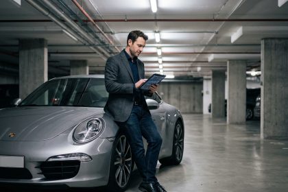 Homme en blazer consultant des documents d'assurance appuyé contre une Porsche 911 argentée dans un parking souterrain